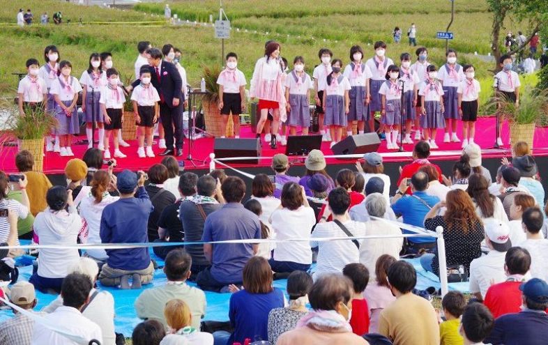 赤米の田を背景に新本小児童と一緒に歌う相川さん（中央）