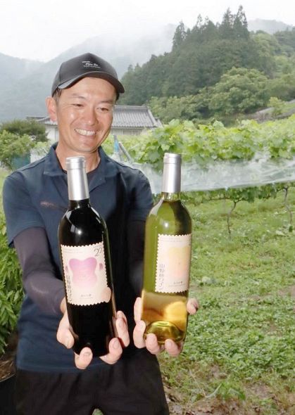 ブドウ畑の前でサミット提供ワインの新酒を手にする峯松さん