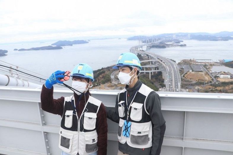 瀬戸大橋の塔頂部からの景色をバックに記念撮影するスカイツアーの参加者。２０２４年は「常設化」が見送られる＝２３年３月