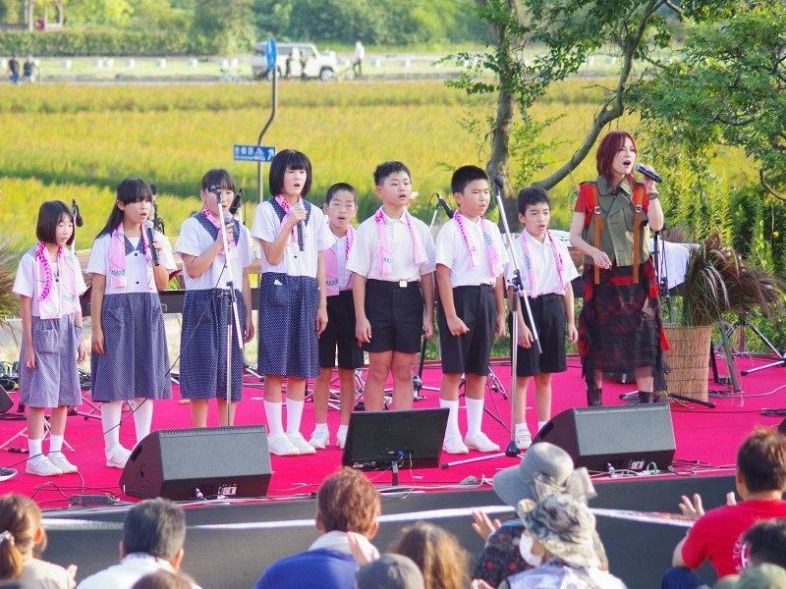 赤米の田を背景に新本小児童と一緒に歌う相川さん（右）